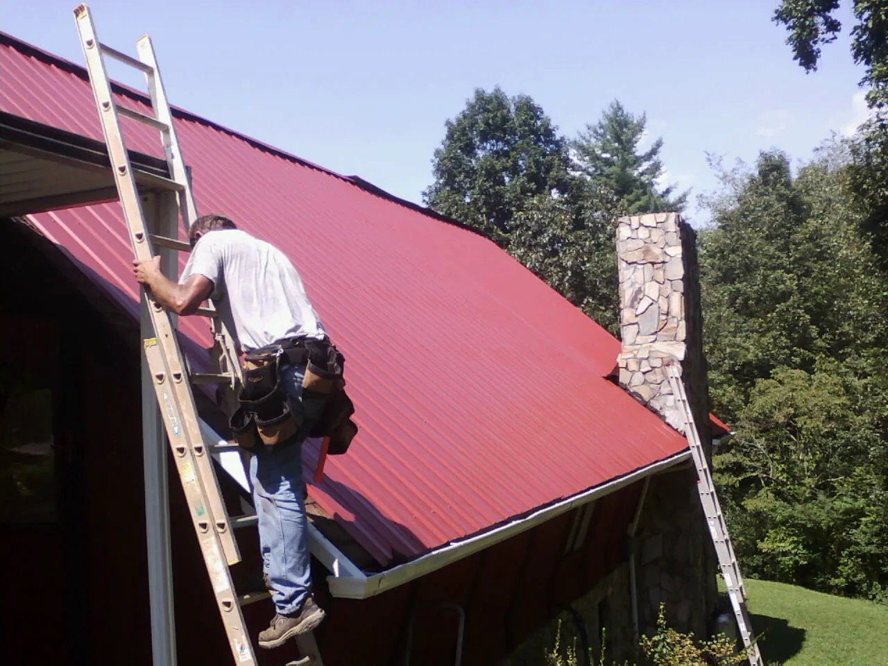Roofer on a ladder beside a red metal roof and stone chimney — Soffit & Fascia Repair by Roofing Brothers in Hauppauge